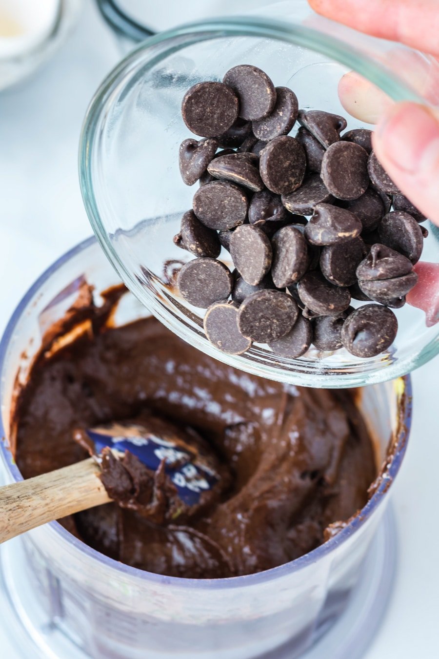 Avacado brownie batter in mixer with a hand holding a bowl of chocolate chips to add in