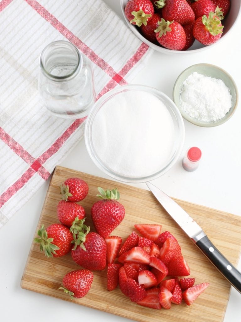 chopped and whole strawberries on a cutting board with knife next to a bowl of cornstarch and sugar