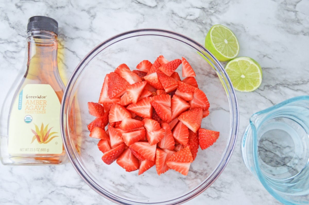 agave sweetener with sliced strawberries in a bowl a sliced lime and water in a pitcher on a counter