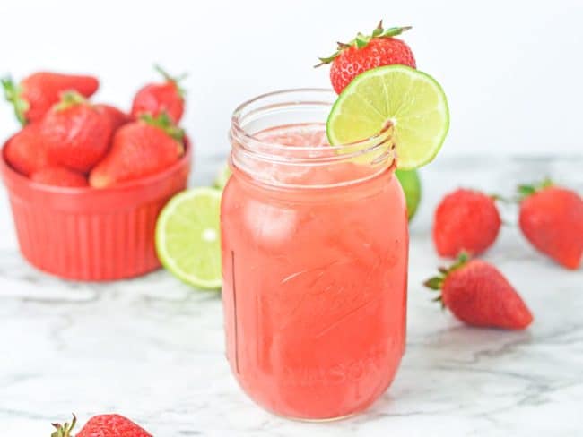 strawberry agua fresca in a mason jar glass with ice and scattered fruit in the background