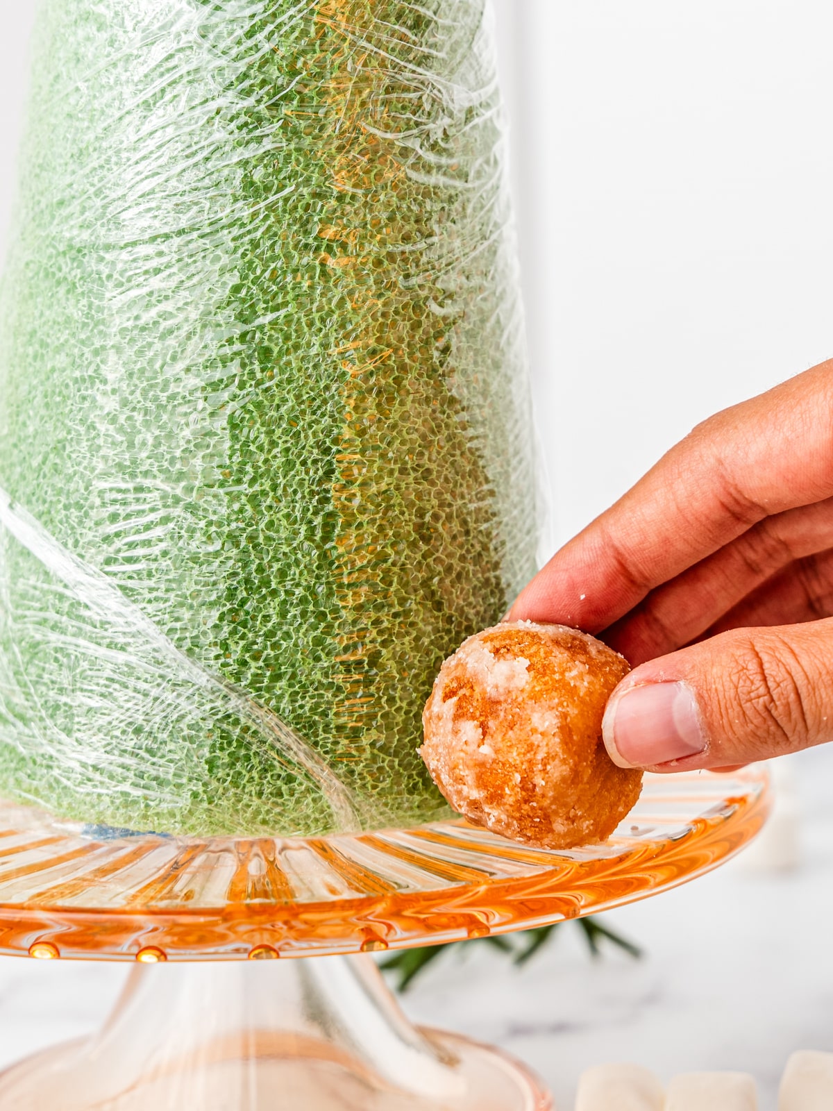 A hand pushing a donut hole onto a toothpick at the base a plastic wrapped styrofoam cone.