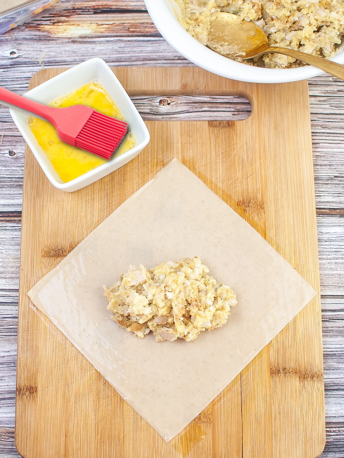 A wooden cutting board with a filled egg roll wrapper next to a bowl of egg wash.