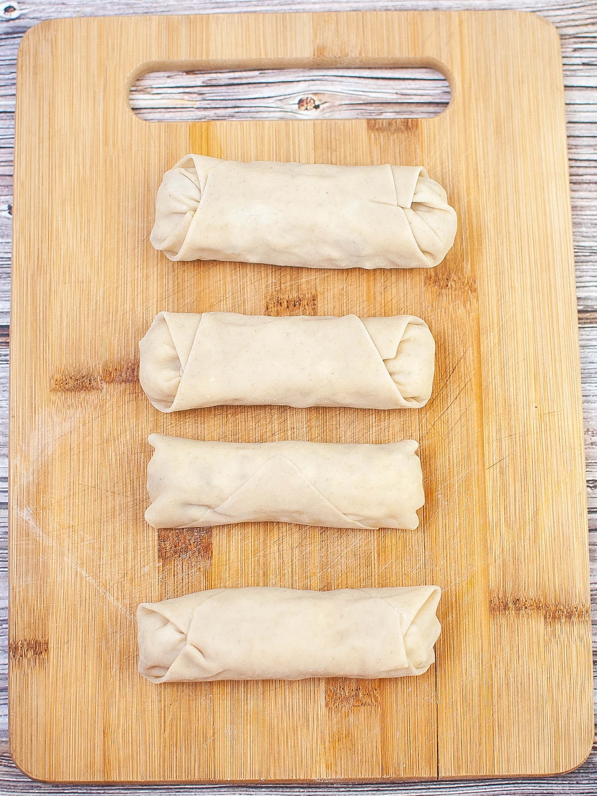 Four uncooked eggrolls on a wooden cutting board.