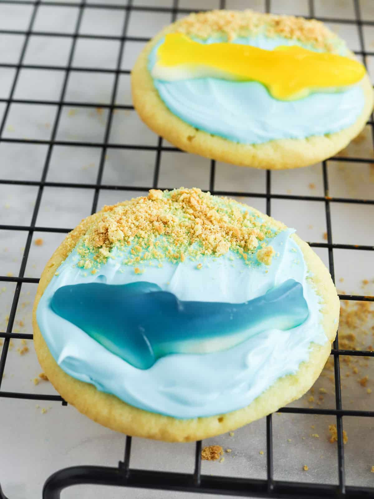 Two cookies decorated with blue and yellow icing on a cooling rack.