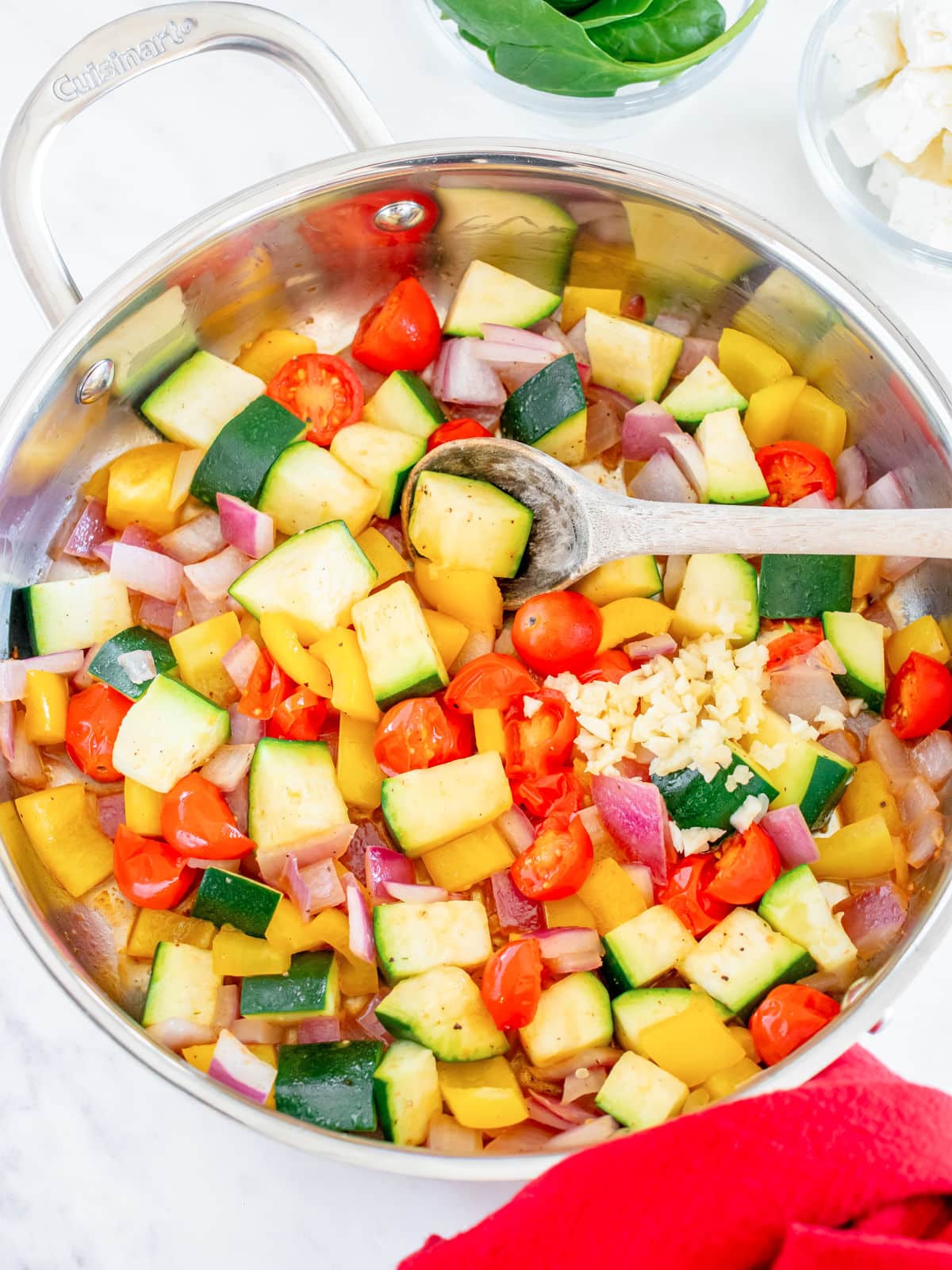 A variety of chopped vegetables including zucchini, tomatoes, bell peppers, and onions in a stainless steel pan with a wooden spoon.