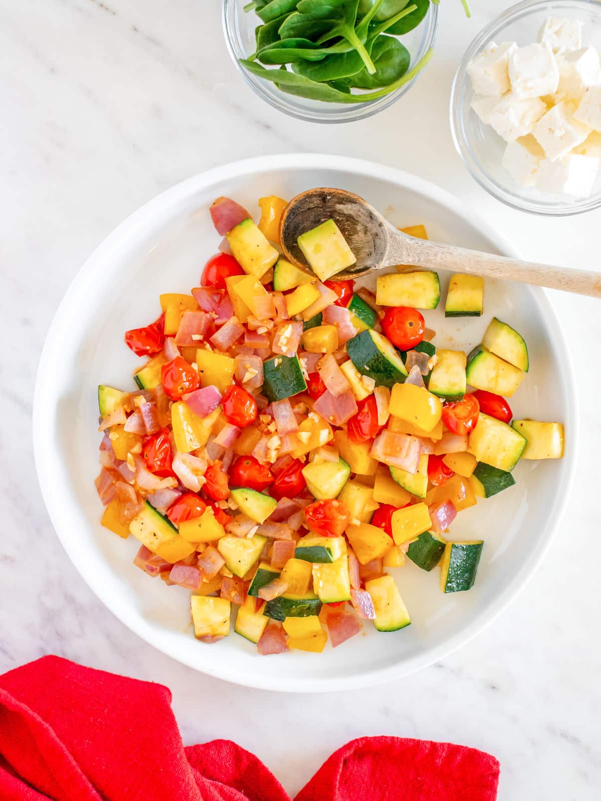 A bowl of fresh diced vegetable salad with zucchini, tomatoes, and yellow bell peppers, accompanied by a side of greens and cheese.