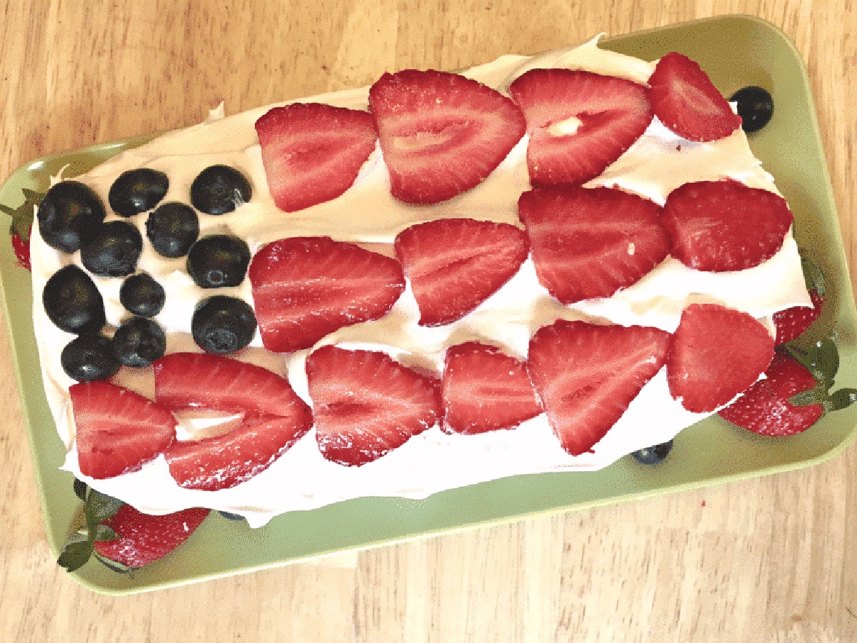 A rectangular cake topped with cream and decorated with sliced strawberries and blueberries on a green tray.