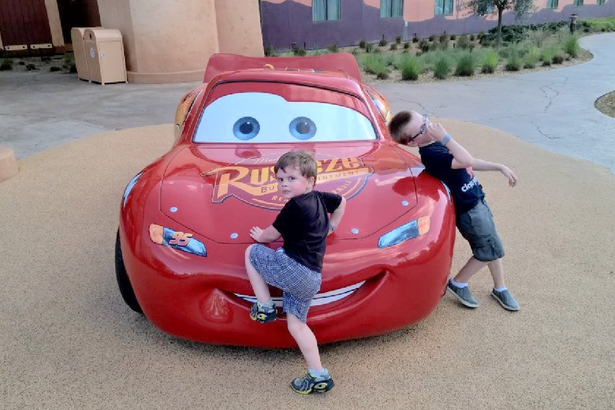 Two boys posing playfully beside a life-sized model of lightning mcqueen from the movie "cars" at a themed park.