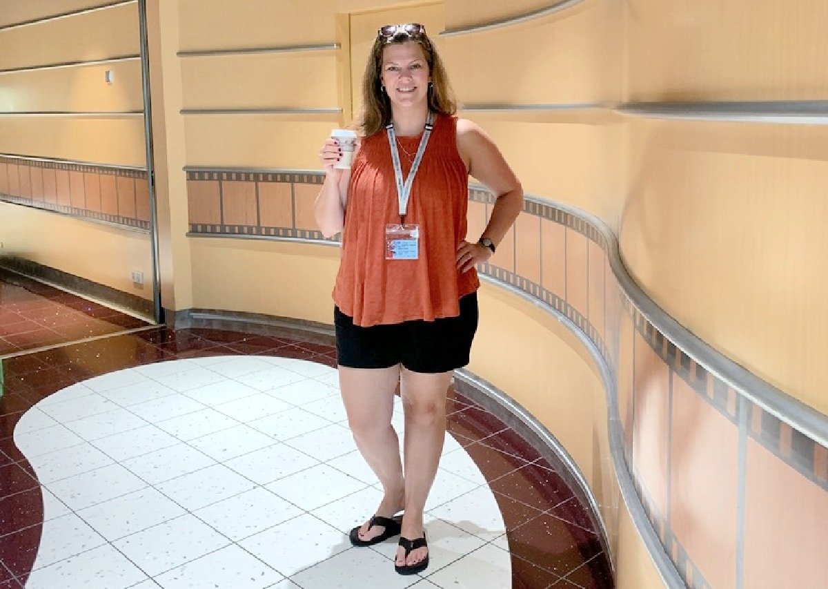 A woman in an orange top and black shorts, holding a coffee cup, smiles while standing in a curved hallway with warm-toned walls.