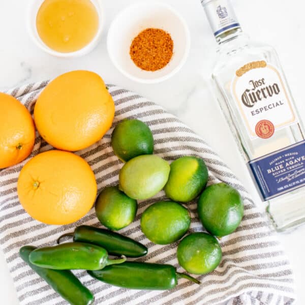 Oranges, limes, jalapeños, a bottle of José Cuervo Especial, and two small bowls containing liquid and spices on a marble countertop for a Cinco de Mayo party.