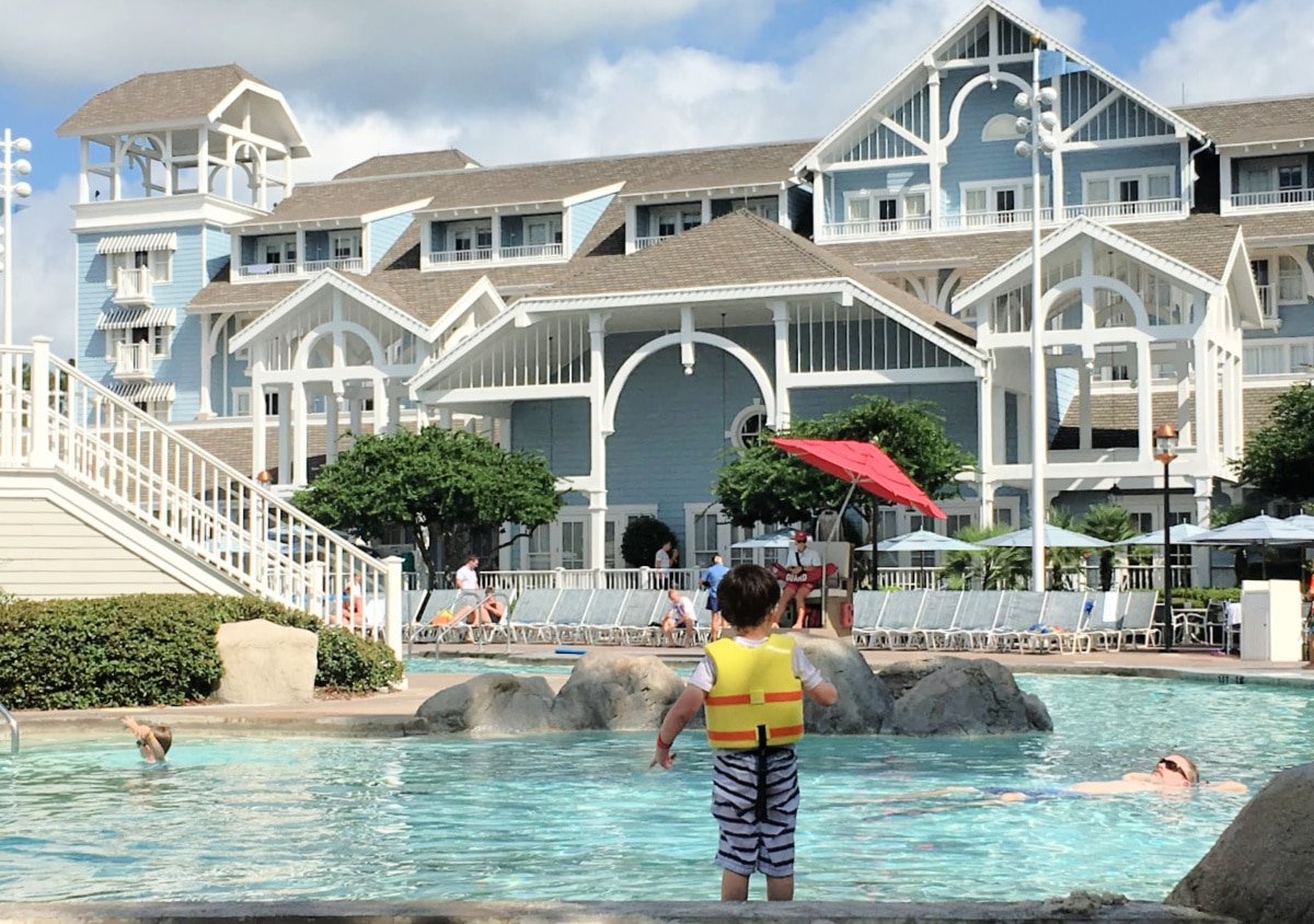A young child in a life jacket stands by a pool at a resort with white, victorian-style architecture and people lounging.