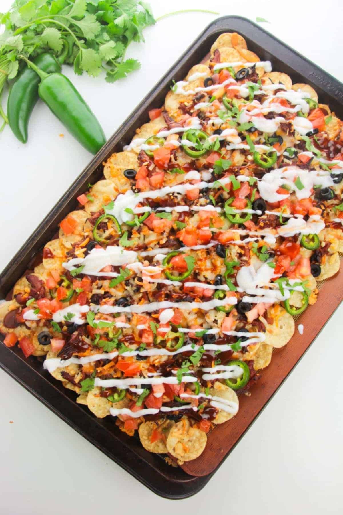 A tray of loaded nachos with jalapeños, tomatoes, black olives, and a drizzle of sour cream on a white background.