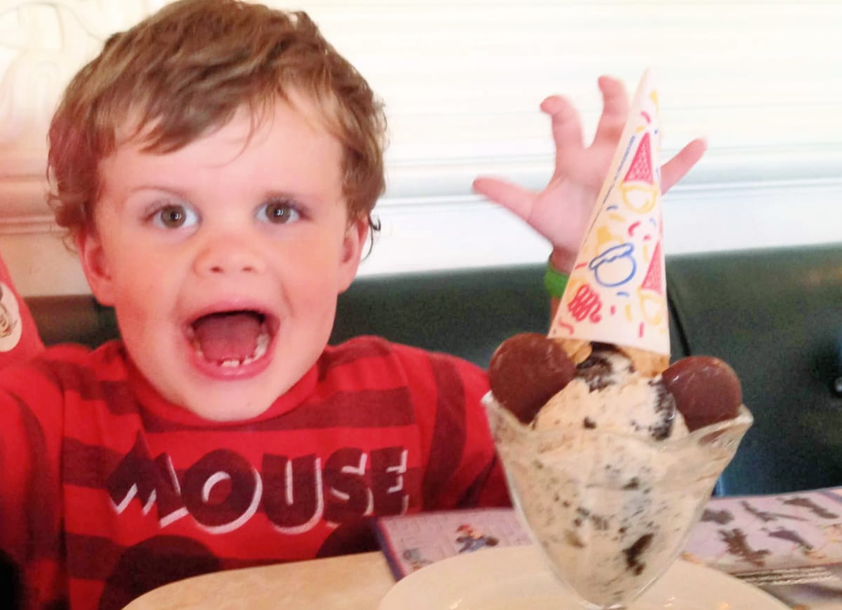 Young boy in a red striped shirt excitedly reaching for a bowl of ice cream with a birthday hat on, sitting at a table.