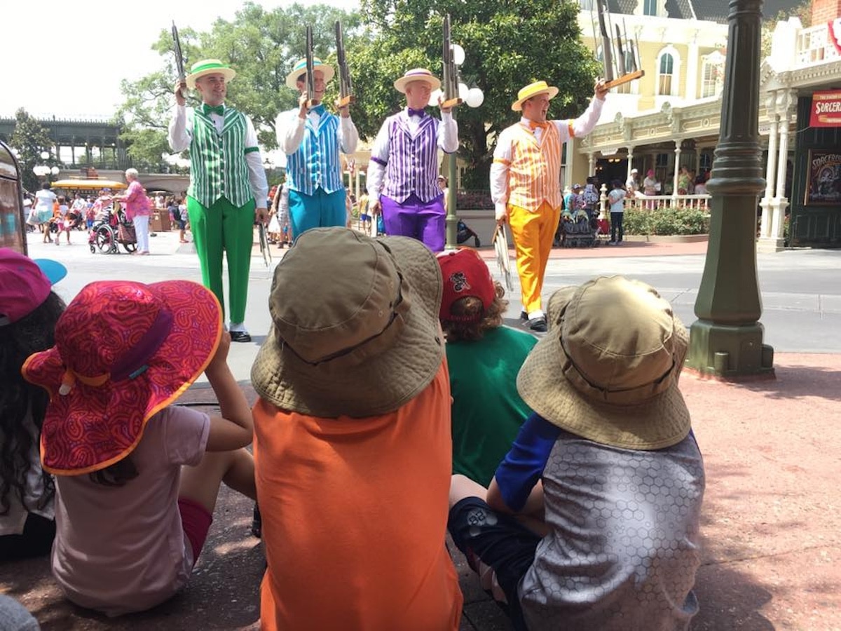 Children in colorful hats watching a barbershop quartet dressed in vibrant striped suits performing on a sunny street.