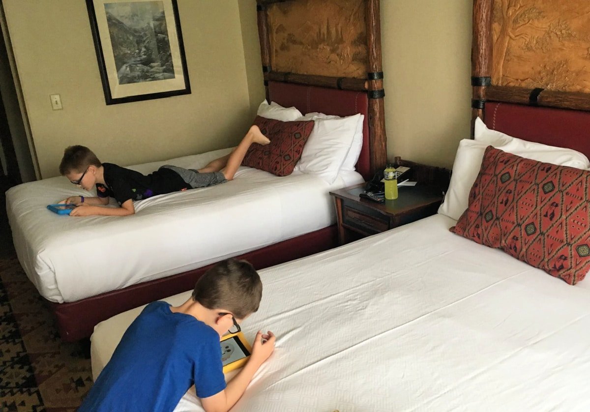 Two boys using tablets while lying on separate beds in a hotel room with patterned rugs and decorative headboards.