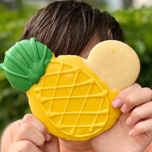 A child holding a pineapple-shaped cookie in front of their face, with a green top and yellow base.