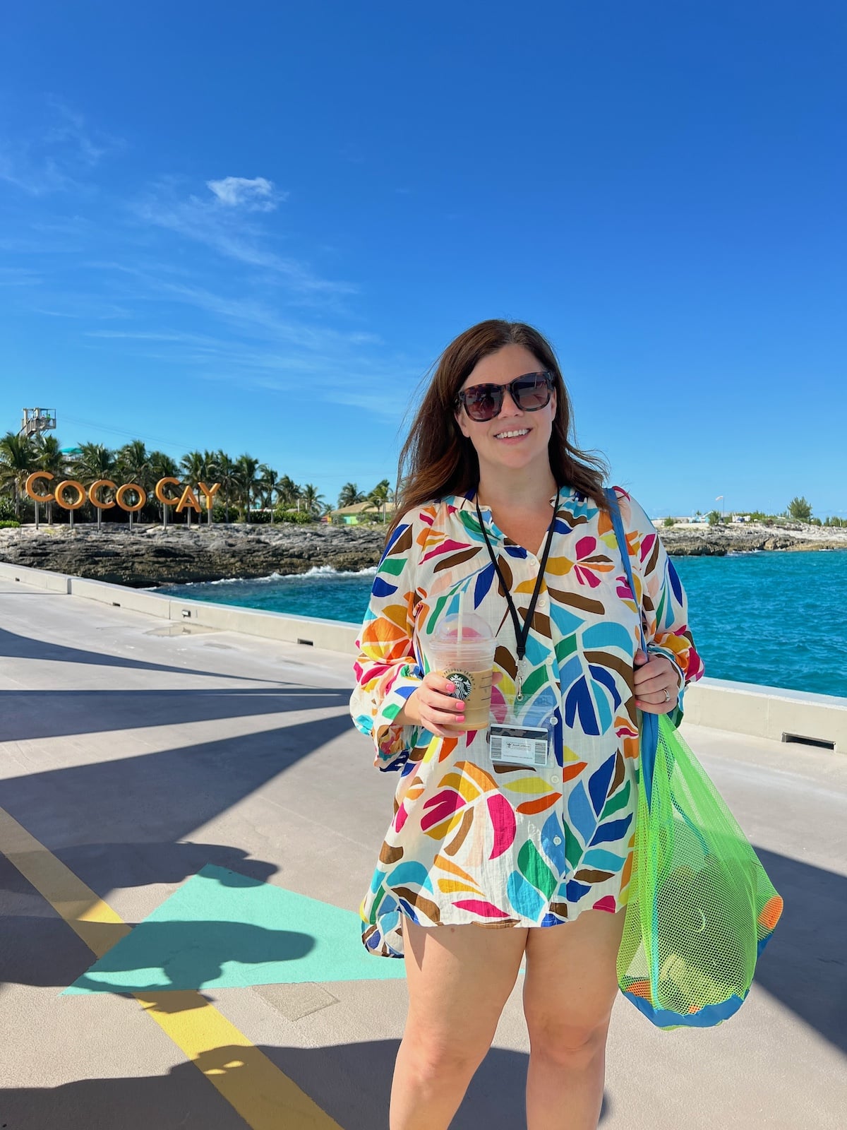 A woman in sunglasses and a colorful dress stands on a sunny boardwalk holding a mesh bag. The background features a sign reading "Coco Cay" and an ocean view.