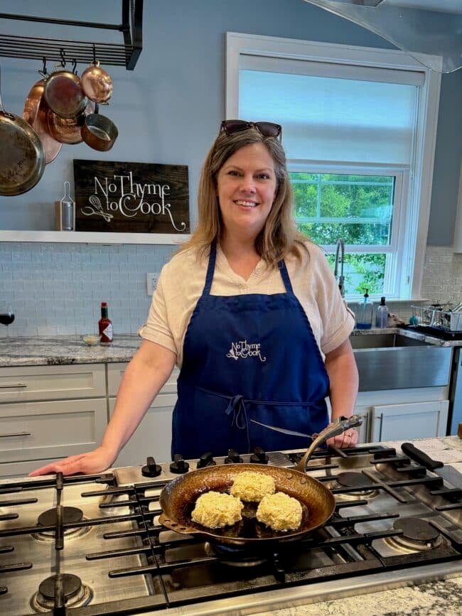 A woman wearing an apron stands in a kitchen and smiles at the camera. She is cooking crab cakes in a skillet on the stovetop. The kitchen has a sign behind her that reads "No Thyme to Cook." About Andrea Updyke, she truly knows how to add a personal touch to every dish.