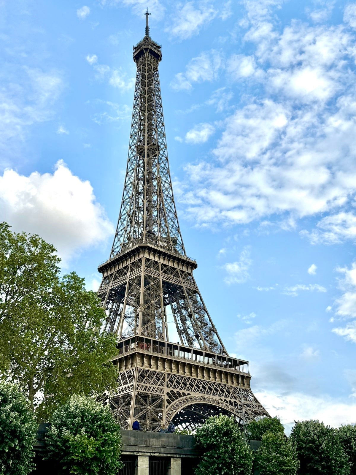 The Eiffel Tower stands tall against a blue sky with scattered clouds, surrounded by green trees at its base, creating an unforgettable sight for anyone spending one day in Paris.