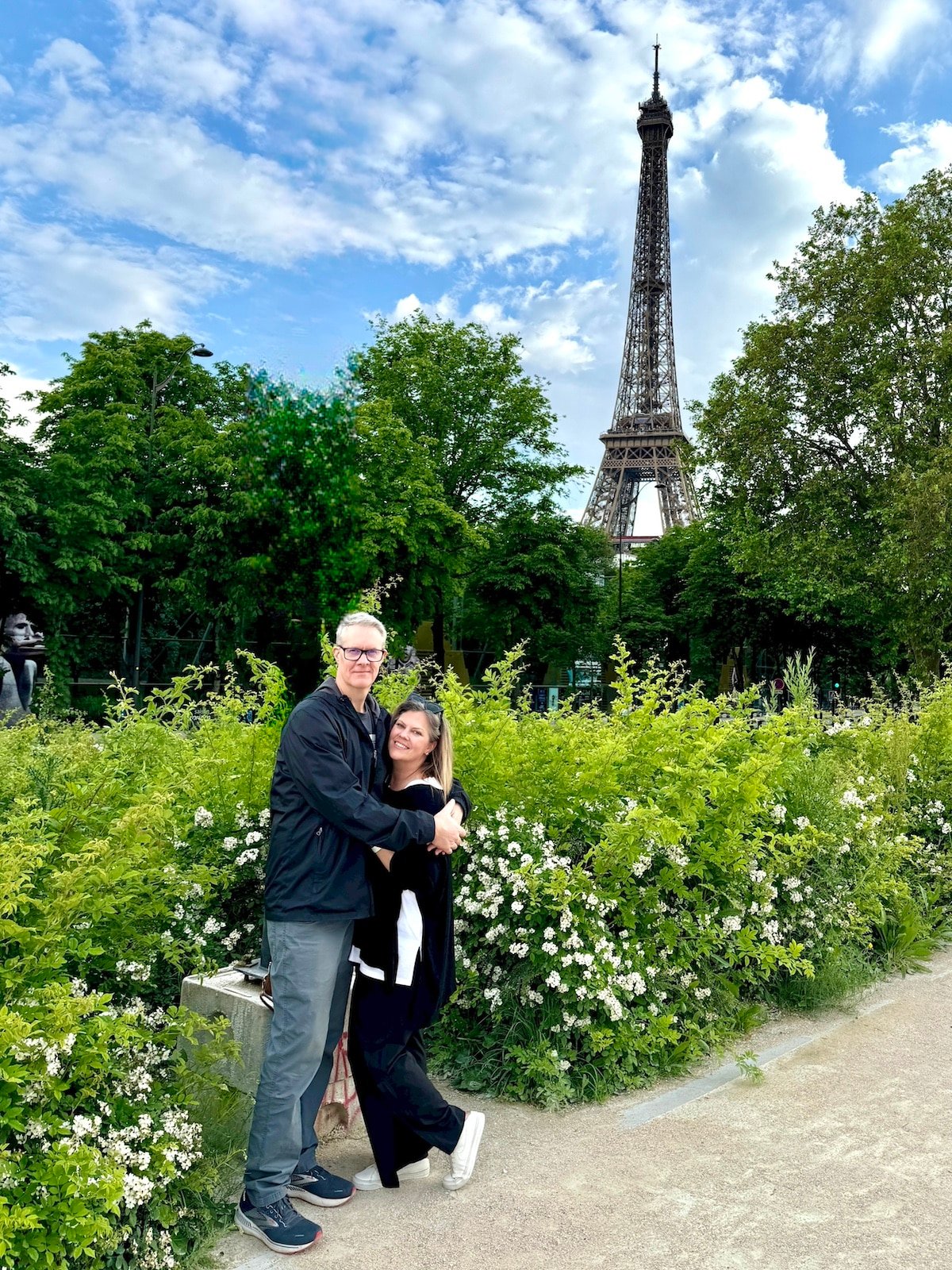 A man and a woman stand together hugging in front of a lush green garden with the Eiffel Tower in the background, capturing their one day in Paris on a clear day.