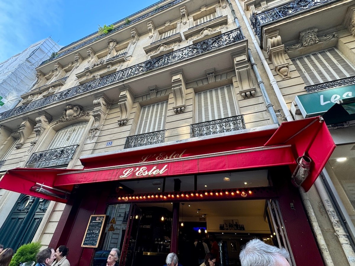Facade of a multi-story building with ornate balconies, shutters, and a red awning reading "L'Estel." People are sitting at tables in front of the building, soaking in the charm as if it were just another day in Paris.