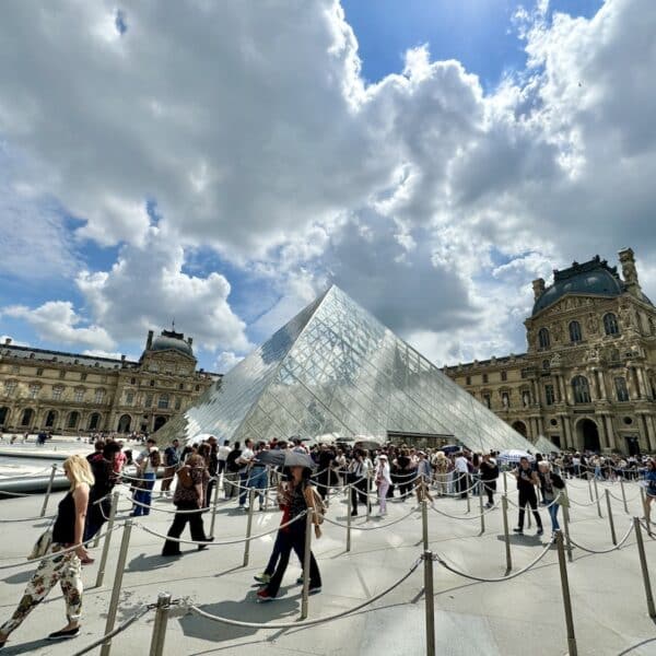 People gather around the glass pyramid entrance of the Louvre Museum in Paris on a sunny day with scattered clouds, making the most of their one day in Paris.