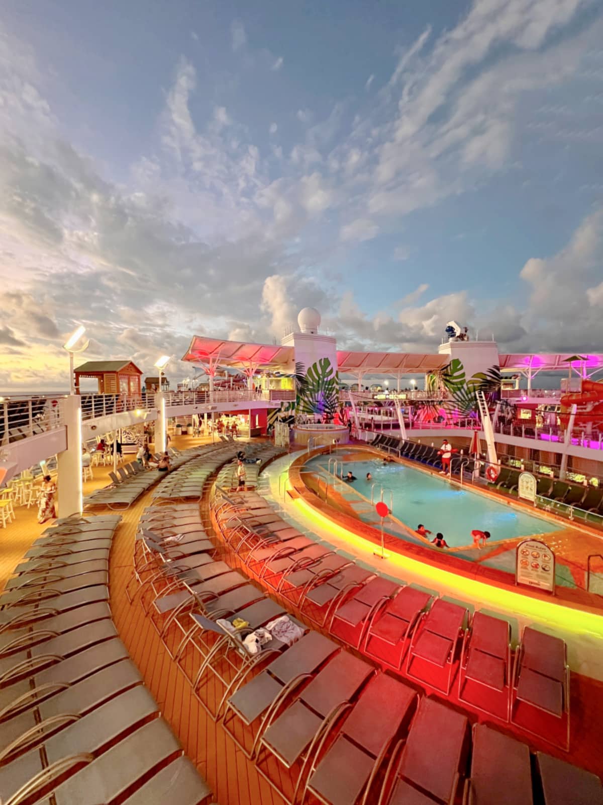 A luxurious cruise ship deck at twilight with rows of lounge chairs surrounding a lit pool. People can be seen swimming and relaxing. The sky is filled with clouds and hues of blue and pink.