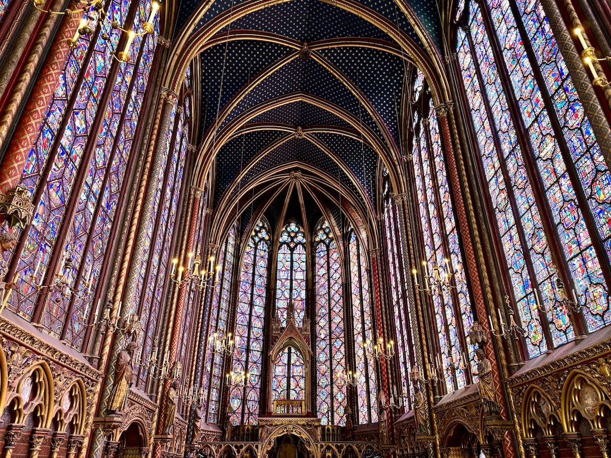 Interior of a Gothic-style chapel featuring high arched ceilings, intricate stained glass windows, ornate carvings, and elaborate chandeliers—reminiscent of what you might see during one day in Paris.