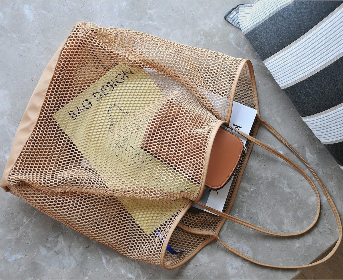 A yellow book and a beige wallet inside a mesh tote bag placed on a marble surface next to a striped cushion.