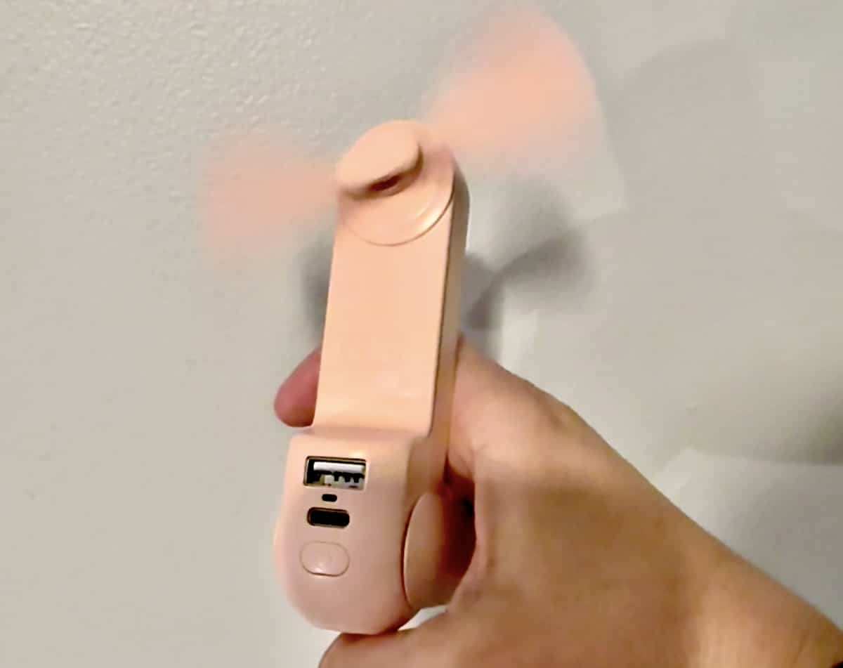 A hand holding a small, pink handheld fan with a USB port and a micro-USB charging port. The fan blades are spinning.
