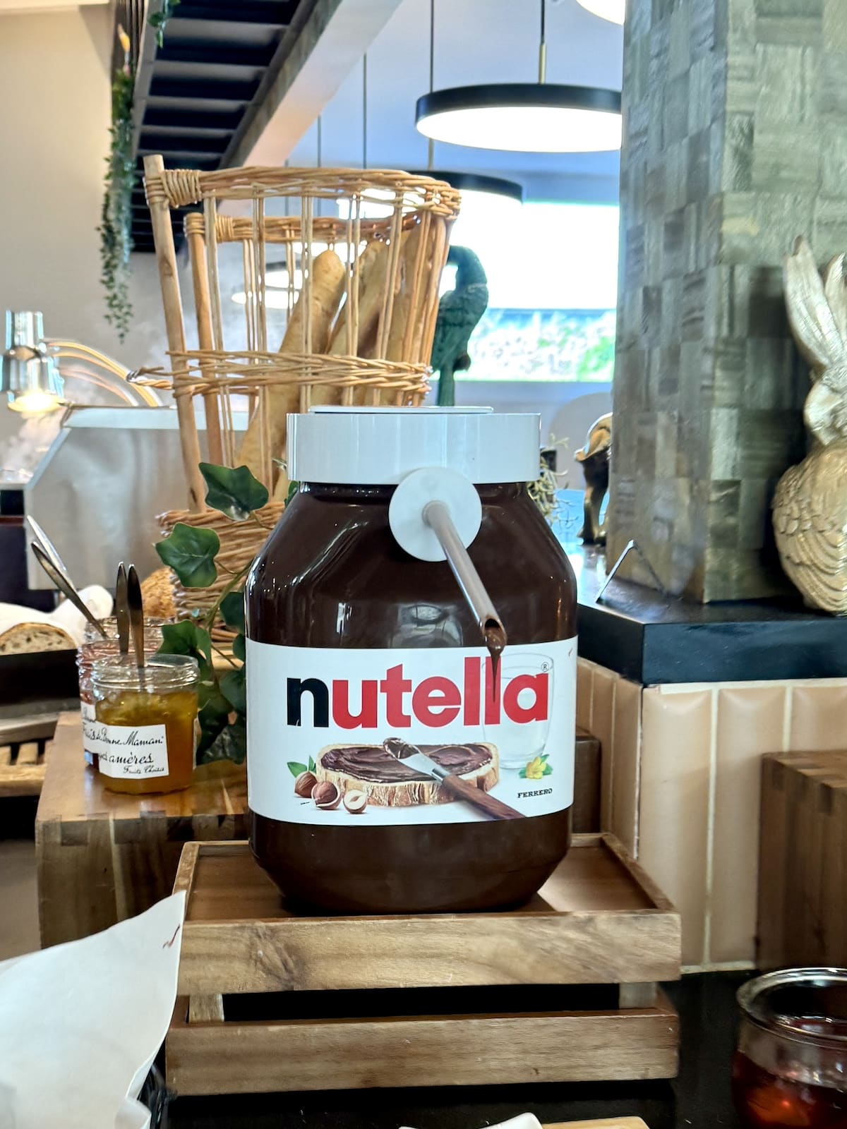 Large Nutella jar with a spoon, displayed on a wooden stand in a kitchen setting reminiscent of one day in Paris. Counter with various condiments and a basket in the background.