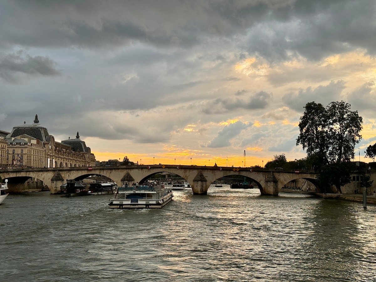 A stone arch bridge spans a river with boats underneath. The sky is cloudy with a hint of sunset, casting a warm glow on the horizon as if capturing one day in Paris. Buildings and trees line the waterfront, completing the picturesque scene.