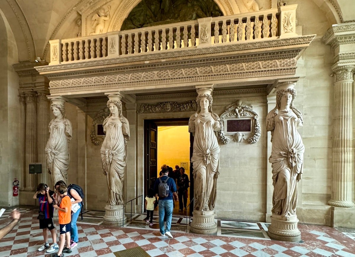 People entering an ornate room decorated with classical statues of women in a grand historical building, reminiscent of something one might experience during an unforgettable day in Paris.