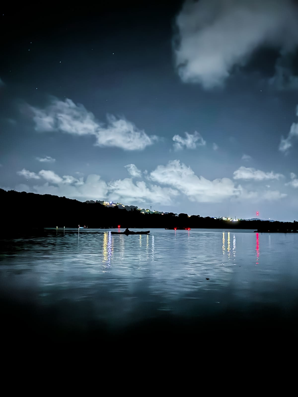 A calm body of water at night reflecting city lights, with distant hills and a partly cloudy sky.