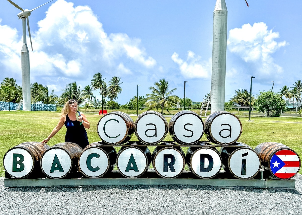 A person stands next to a sign made from barrels reading "Casa Bacardi," with wind turbines and palm trees in the background—one of the must-see things to do in Puerto Rico.