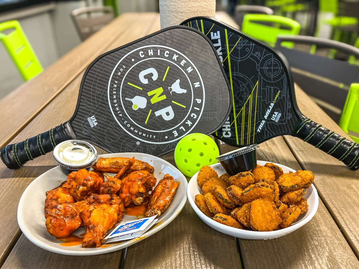 A table with crispy chicken wings, fried pickles, a cup of dipping sauce, a yellow pickleball, and two pickleball paddles displaying the "Chicken N Pickle" logo.