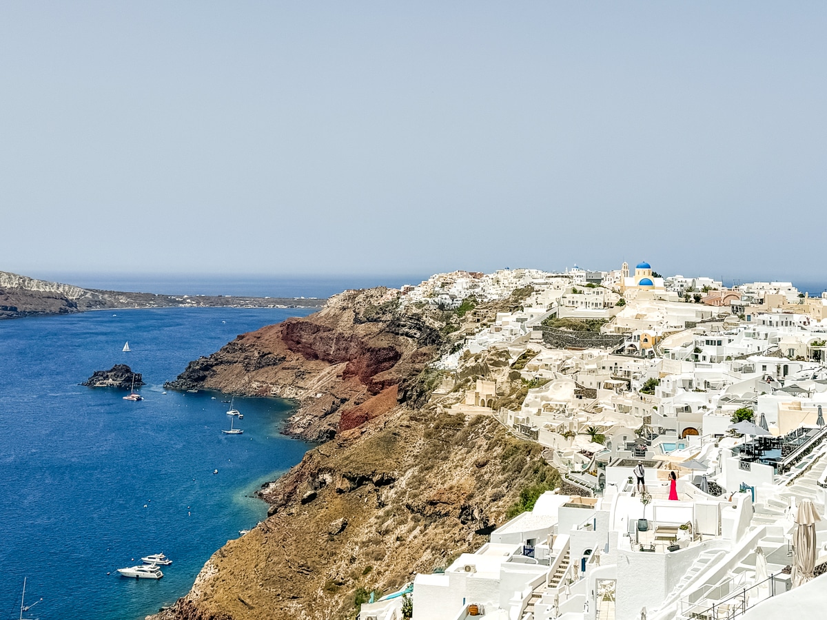 A coastal village with white buildings on a hill overlooking a deep blue sea. There are boats in the water and rocky terrain with white houses spread across the landscape.