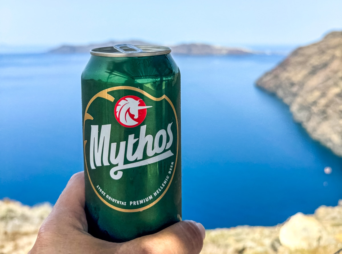 A hand holding a green can of Mythos beer with a scenic view of the coastline and blue ocean in the background.