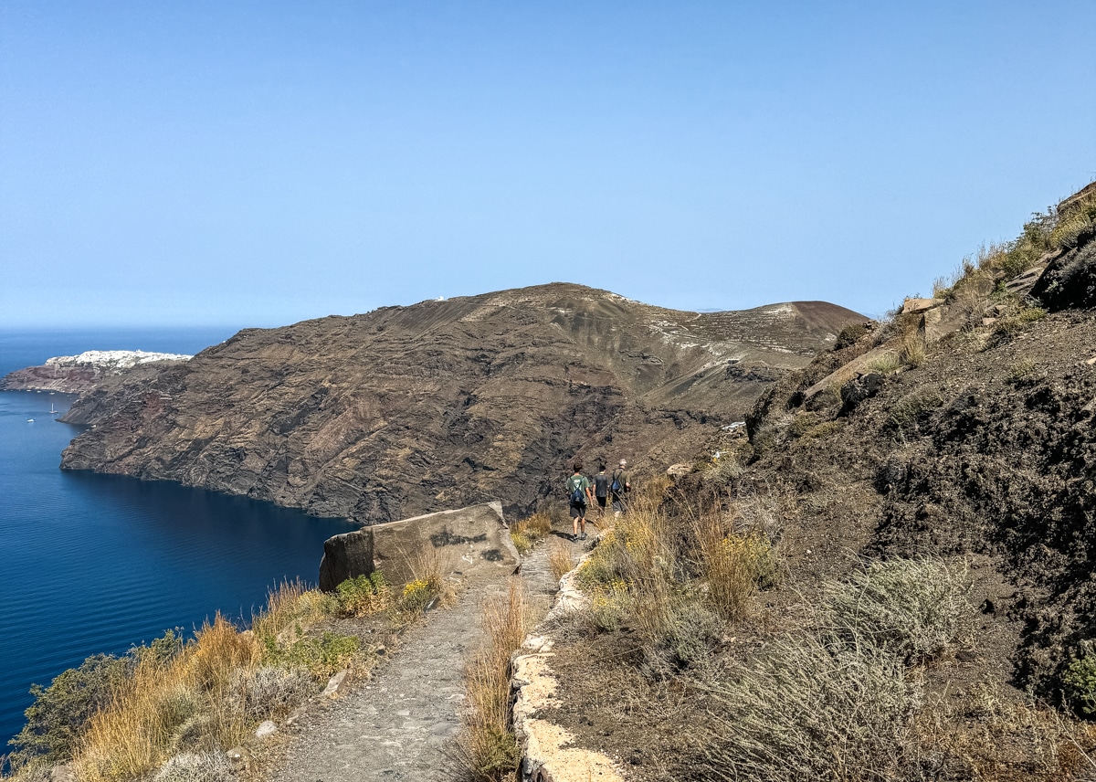 People hiking on a narrow trail along a rugged coastline with steep cliffs, overlooking blue ocean water on a sunny day.