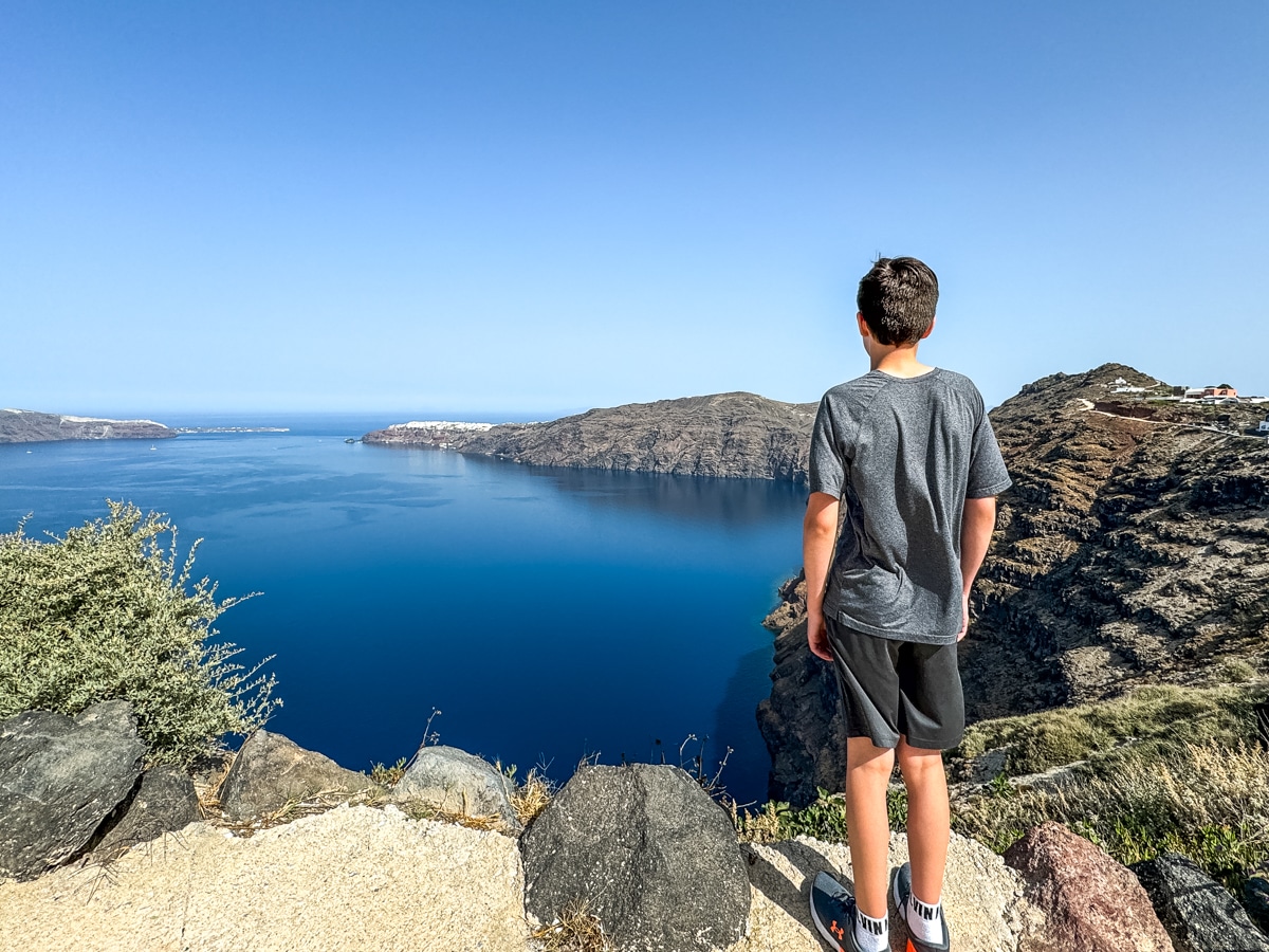 Person in gray shirt and black shorts stands on a rocky cliff, gazing at a vast blue body of water and distant rugged terrain under a clear blue sky.