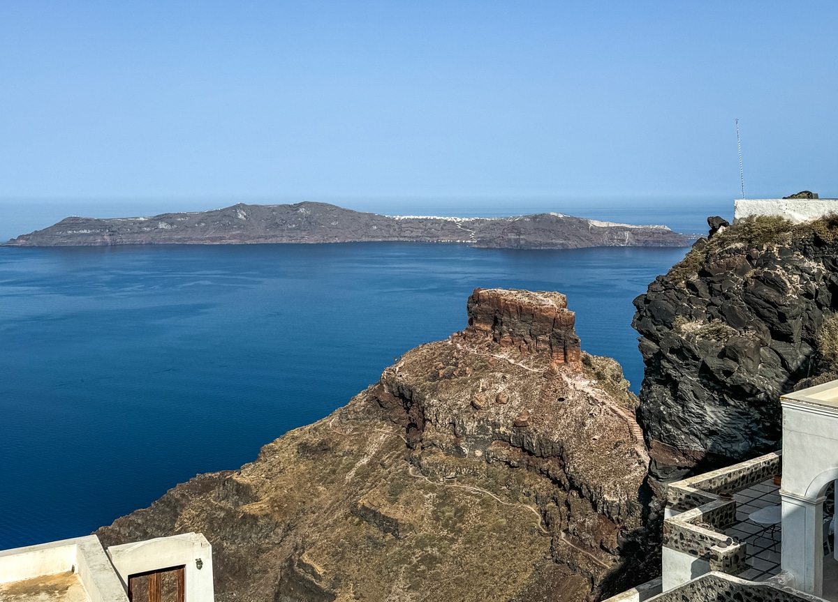 A rugged cliffside overlooking a calm, deep blue sea. In the background, a distant island is visible under a clear blue sky.