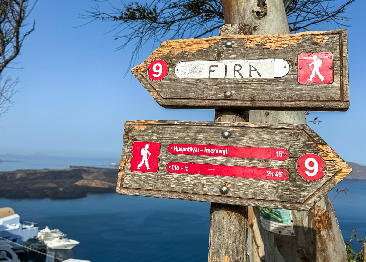 Wooden signs on a tree indicate hiking directions to Fira, Imerovigli (15 minutes), and Oia (2 hours 45 minutes), with a view of the sea and landscape in the background.