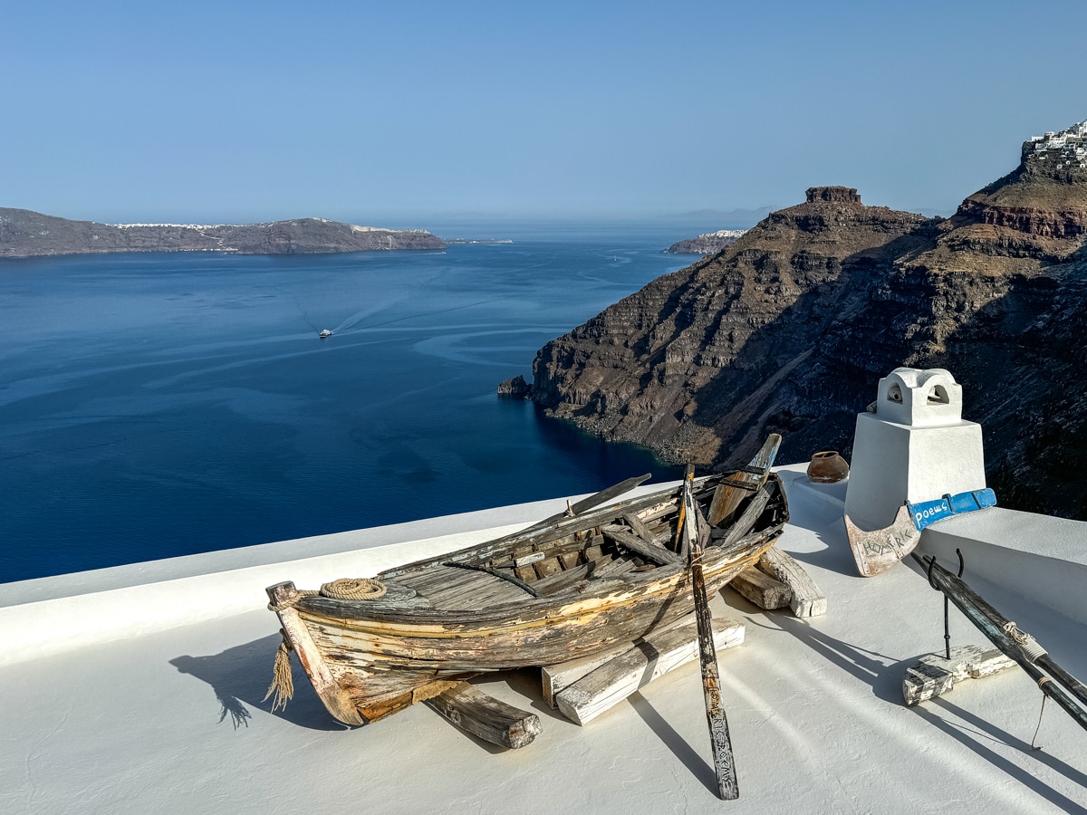 A weathered wooden boat is displayed on a white rooftop overlooking the blue sea and rocky cliffs, with distant land and a clear sky in the background.