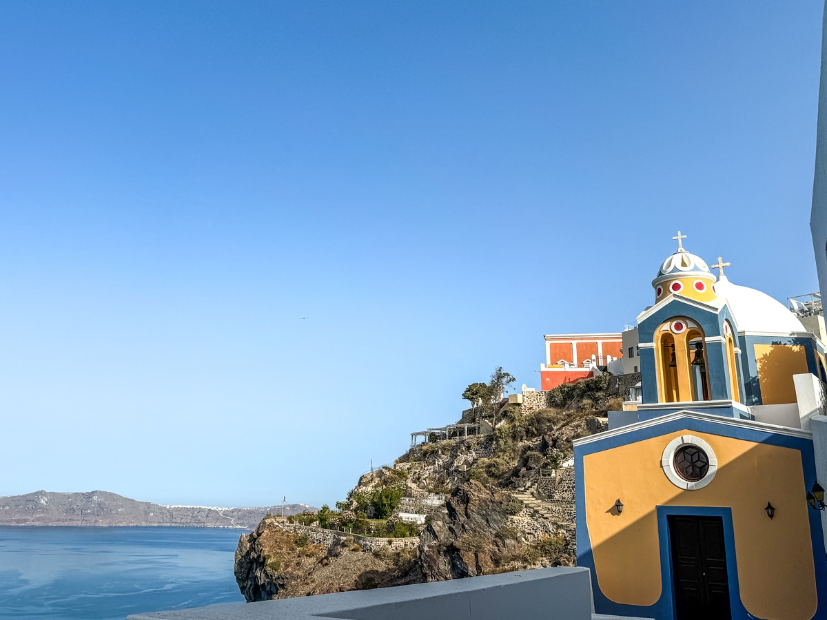 A small, colorful church with dome and crosses stands on a cliffside overlooking the sea, with clear blue sky and distant land in the background.