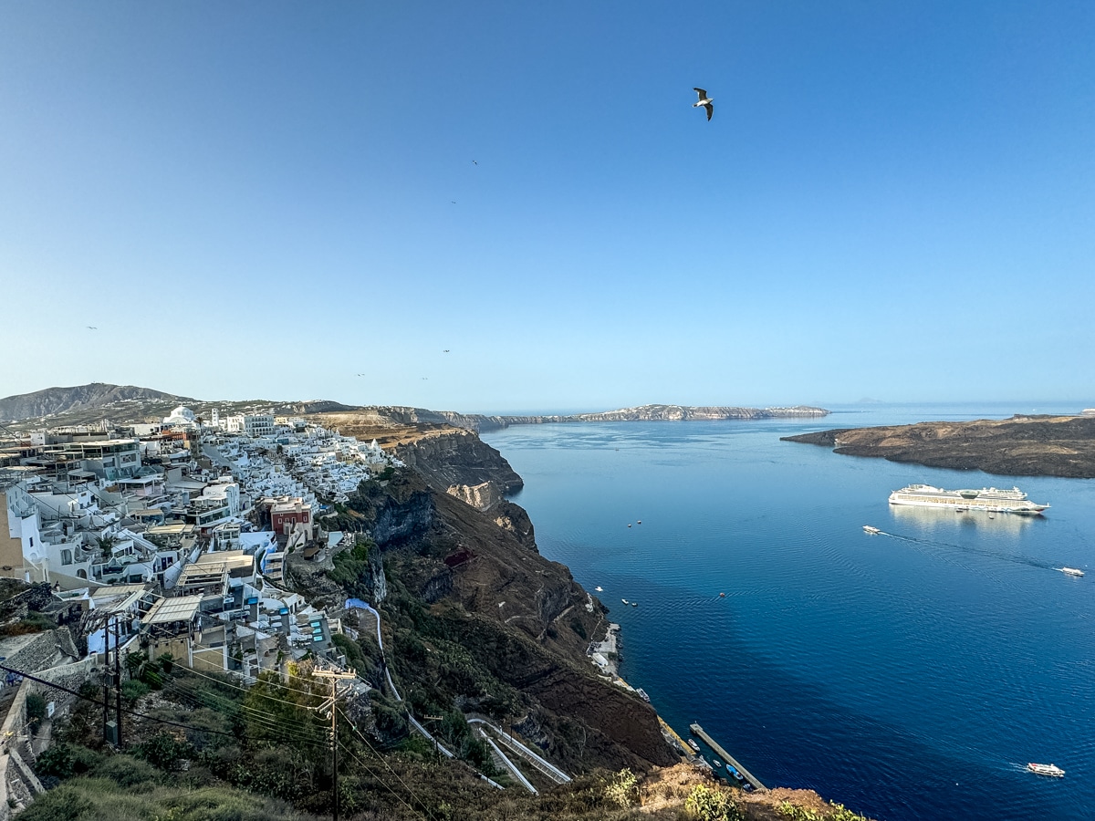 A coastal town with white buildings on a cliff overlooks a calm blue sea with a cruise ship and distant islands; a bird is flying in the clear sky.