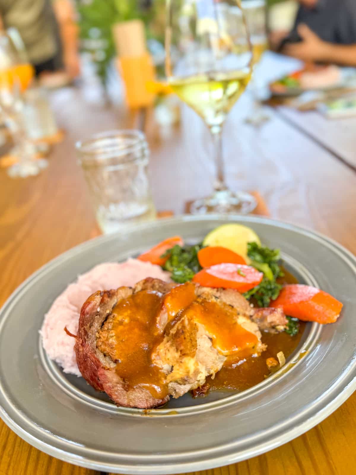 A plate with meatloaf topped with brown sauce, mashed potatoes, sautéed greens, and sliced carrots—just the comfort food you might crave after exploring things to do in Puerto Rico. A glass of white wine sits in the background on a wooden table.