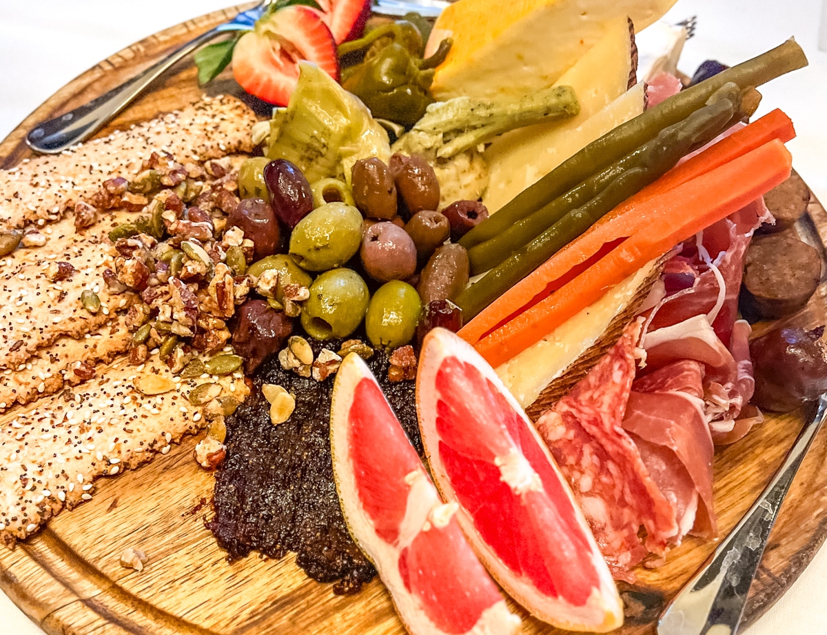 A wooden platter with assorted olives, crackers, nuts, vegetables, cured meats, cheese, and grapefruit slices.