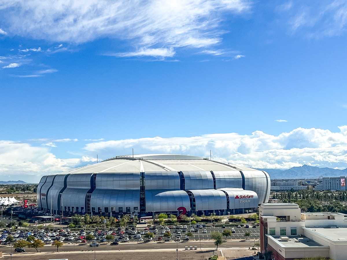 A large, modern stadium with a silver exterior, located in an open area with a partly cloudy sky and mountainous background. Cars are parked in the parking lots surrounding the stadium.