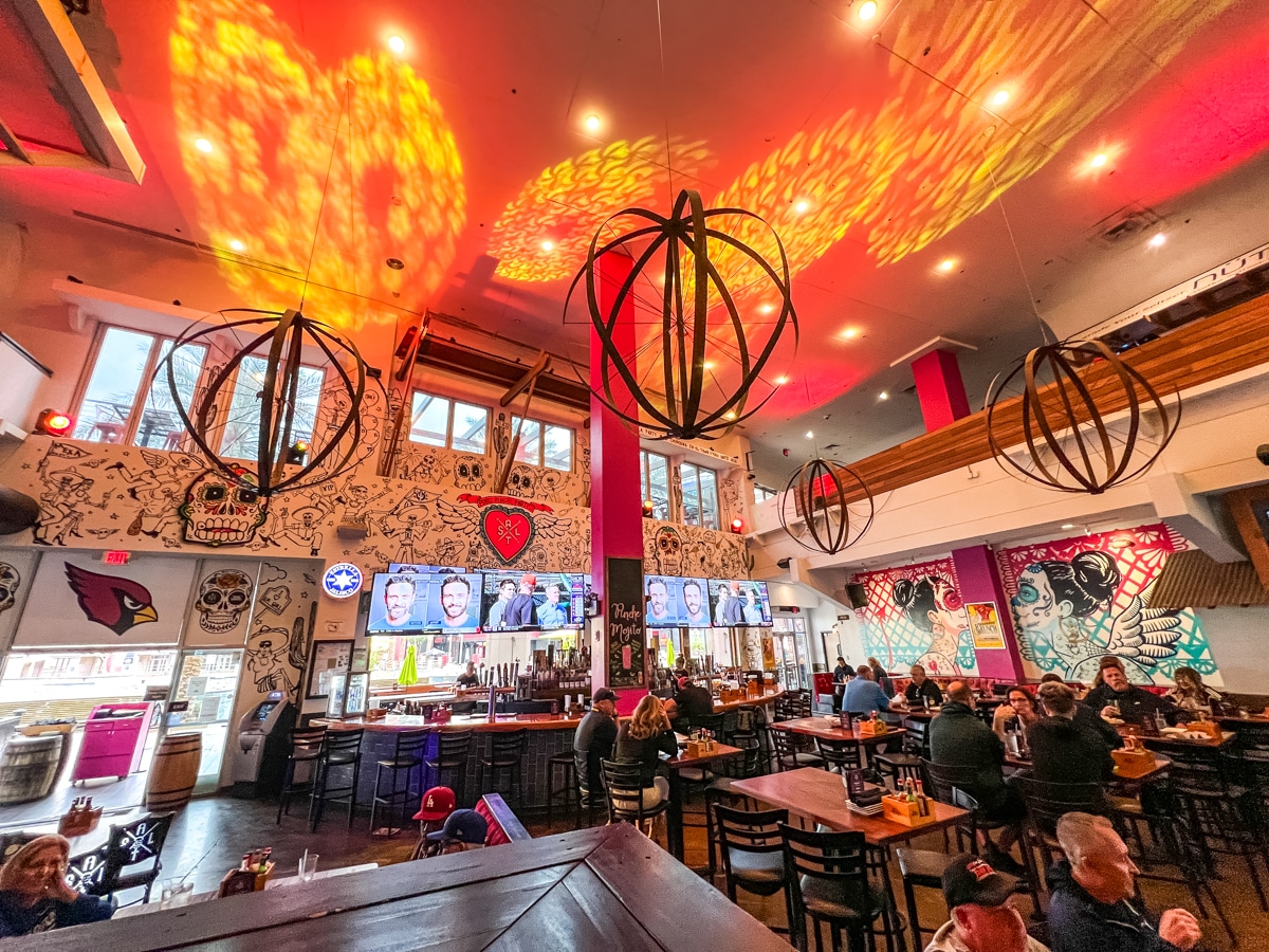 Interior of a brightly lit restaurant with eclectic wall art, modern spherical light fixtures, multiple TV screens above the bar, and patrons seated at tables and barstools.