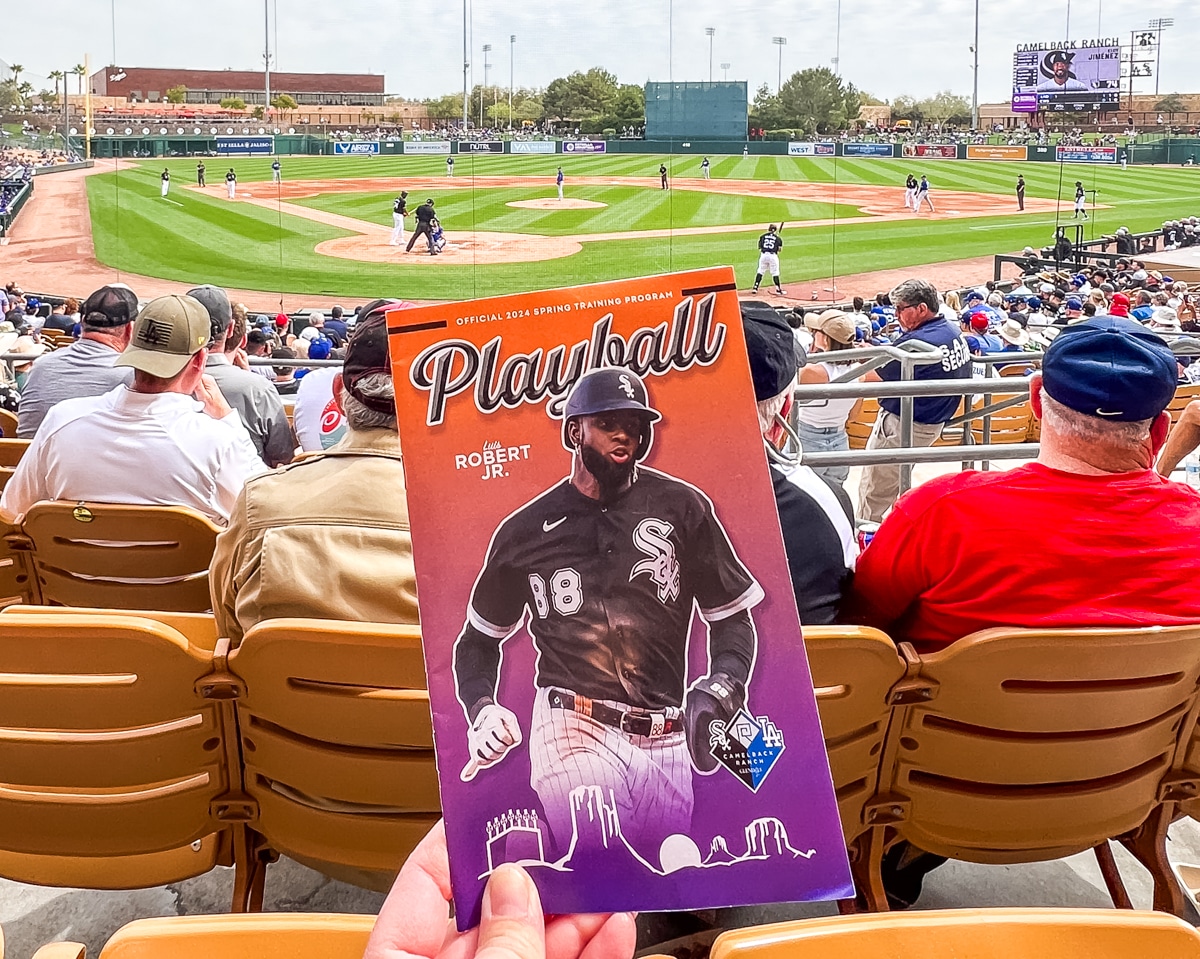 A hand holds a 2023 spring training program at a baseball game. The program features a player in a black uniform. Spectators are seated in the stands while players are on the field.